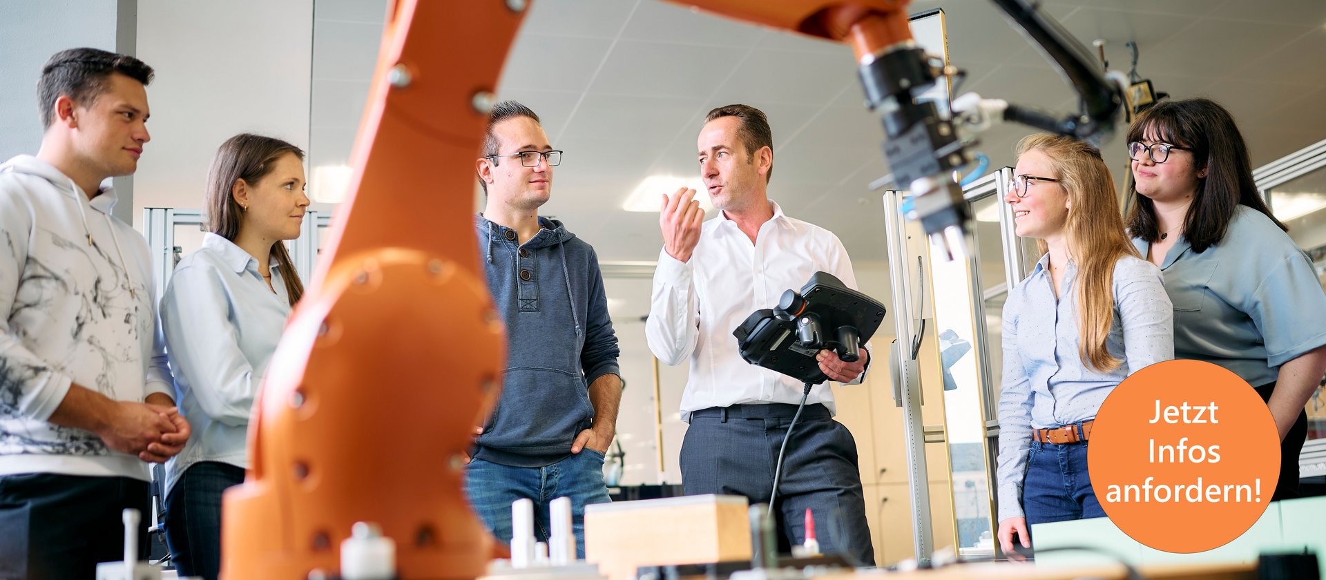 Studierende stehen um einen großen Industrieroboter. Der Professor erklärt etwas und hält dabei ein Steuergerät für den Roboter in der Hand. Der Roboterarm ist im Vordergrund sichtbar und deutet auf eine moderne technische Umgebung hin.