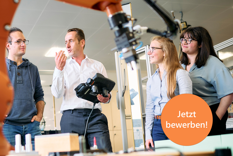 Studierende stehen um einen großen Industrieroboter. Der Professor erklärt etwas und hält dabei ein Steuergerät für den Roboter in der Hand. Der Roboterarm ist im Vordergrund sichtbar und deutet auf eine moderne technische Umgebung hin.